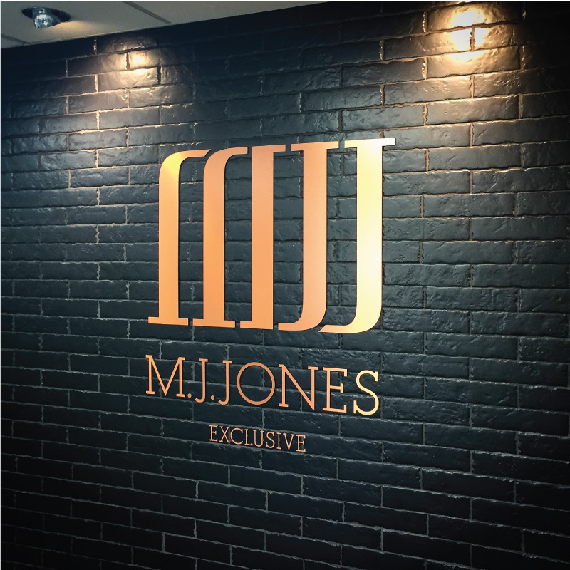 MJ Jones logo in flat cut bronze letters applied to a black brick wall.