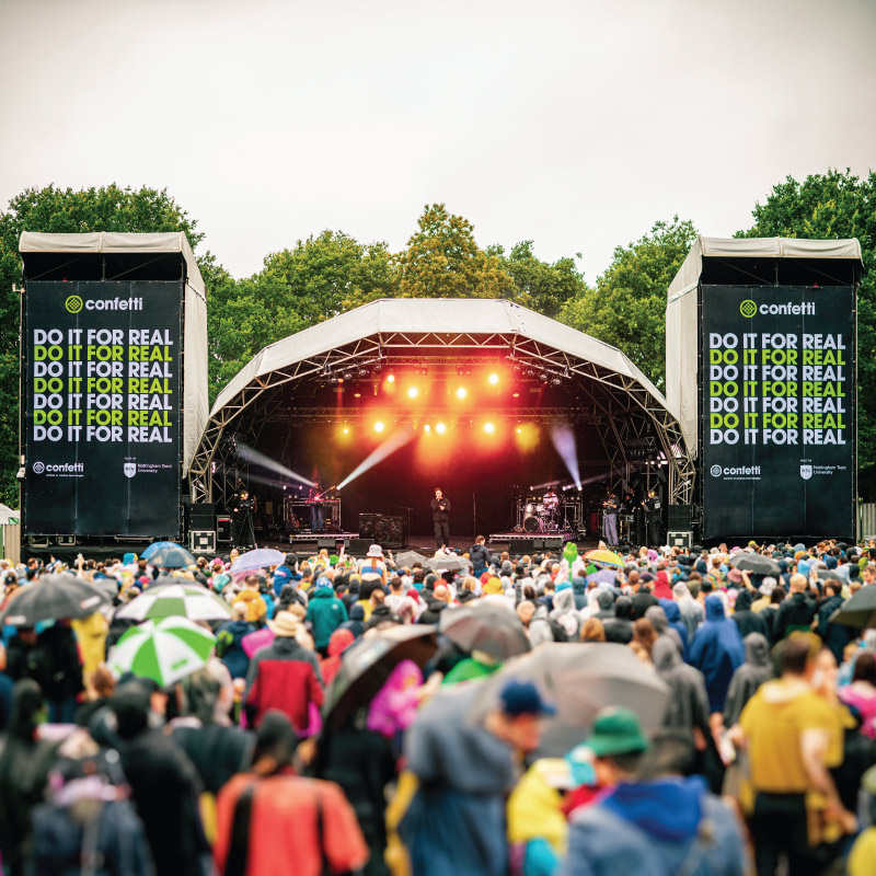 The picture is taken from within the crowd. It shows the Confetti Stage at Splendour festival.Either side of the stage are tall speaker towers. The whole front of these towers have large format banners, they are black with 'do it for real' repeated in white and green. They also show the confetti and nottingham trent university logos.