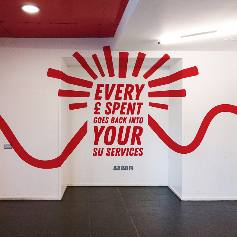 Written in red on a white wall 'Every pound Spent goes back into your SU Services' with red lines in an arch over the top.