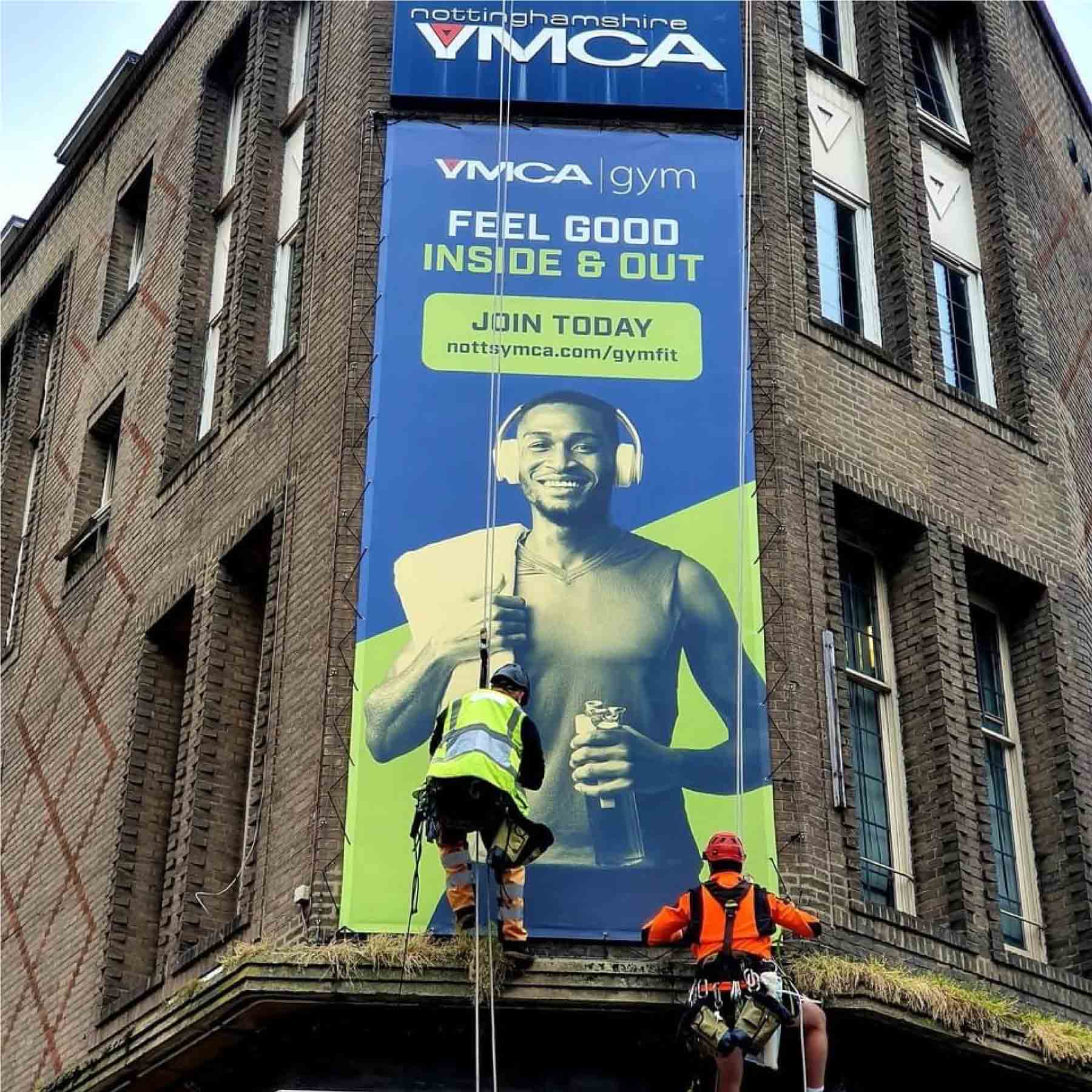 A large scale vertical banner on the corner of a brick building. The banner says 'Feel good inside and out, join today' with a blue background and green accents.
