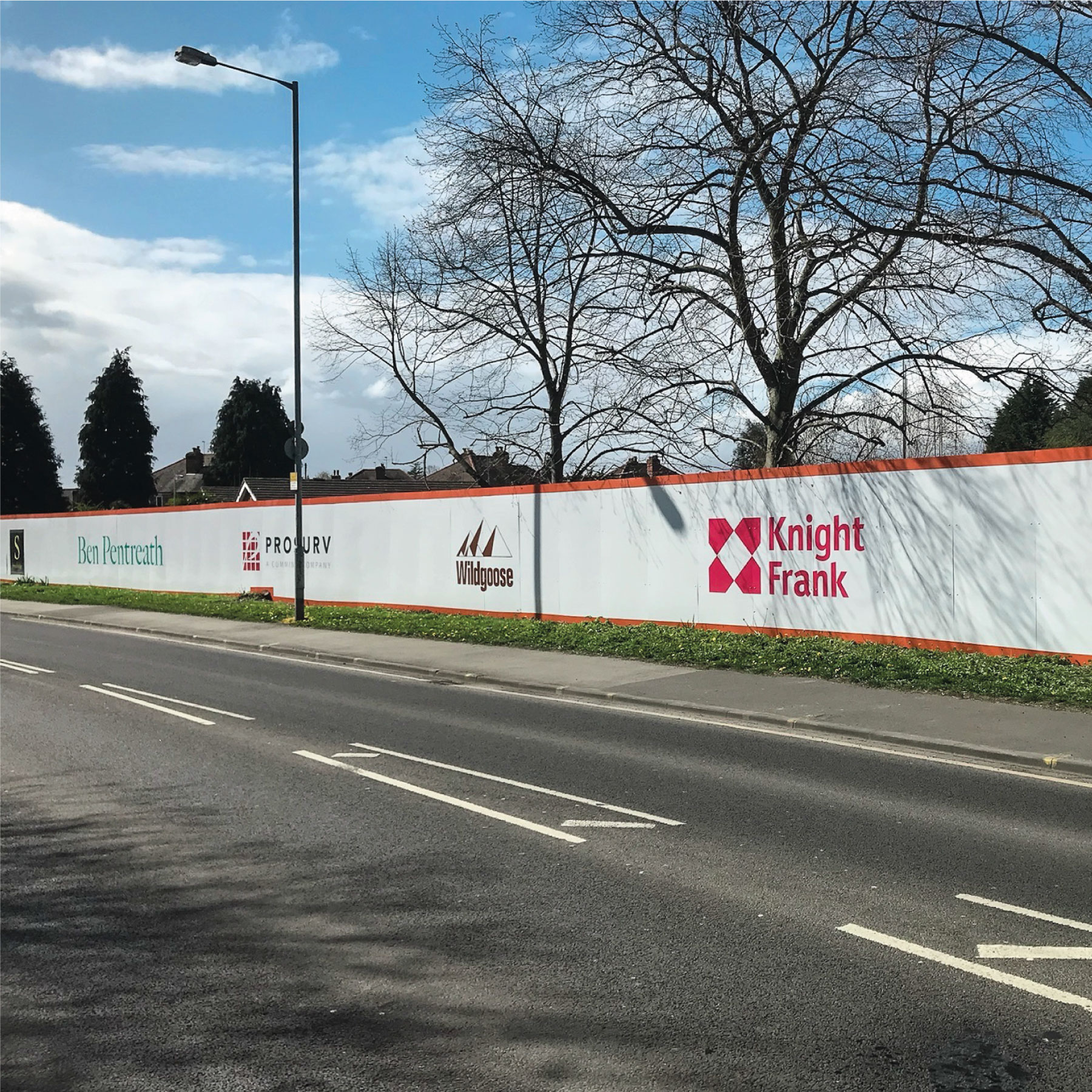 A street lined with solid hoarding boards. The boards have various logos on them and cover the construction site behind.